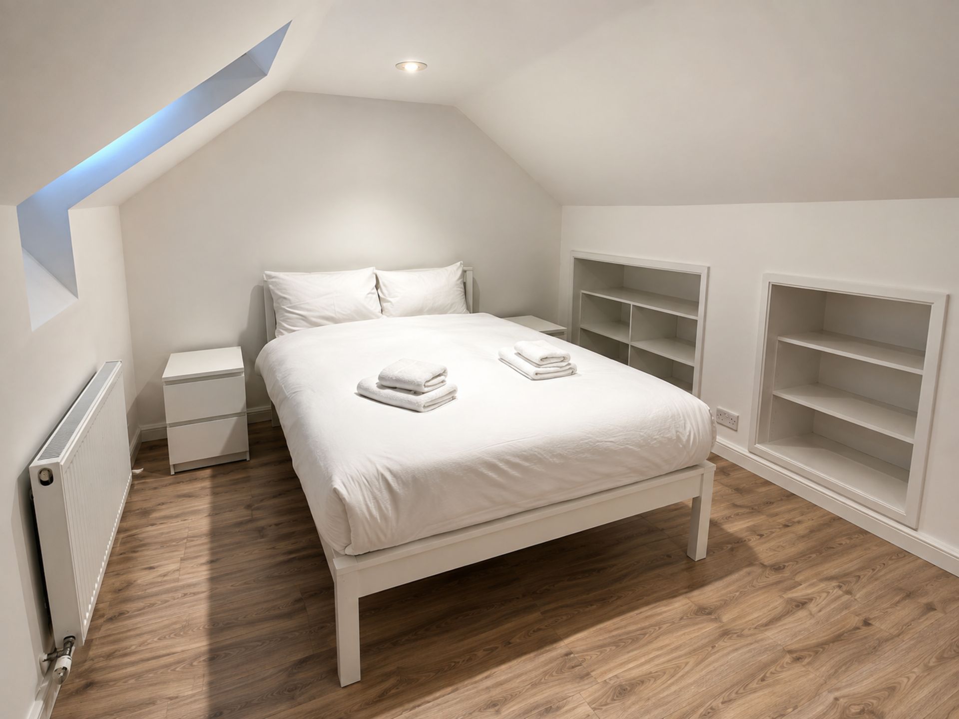 Een slaapkamer met een schuine kap, een bed met witte lakens, een nachtkastje, een radiator en inbouwplanken.