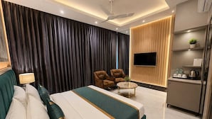 A modern hotel room with a large bed, a seating area with two chairs, a small table, a flat-screen TV mounted on a wooden panel, and a kitchenette area with a coffee maker and water bottles.