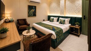 A modern hotel room with a large bed, a desk, two chairs, a small table, an air conditioner, a ceiling fan, and a door.