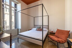 Ein Schlafzimmer mit einem Himmelbett, einem Holzstuhl, einem kleinen Tisch und Blick auf die Stadt durch große Fenster.