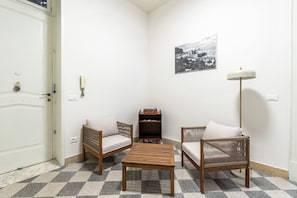 Ein Eckzimmer mit Schachbrettfußboden, zwei Korbstühle mit Kissen, ein kleiner Holztisch, ein Bücherregal, eine Wandlampe und eine Tür mit Griff.