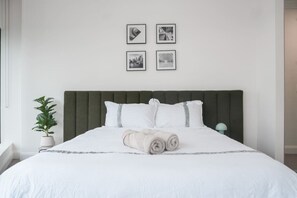 Ein ordentlich bezogenes Bett mit einem grünen Kopfteil, weißer Bettwäsche und einem aufgerollten Handtuch in der Mitte. Darüber an der Wand hängen vier gerahmte Bilder.