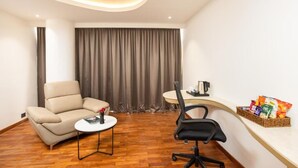 A modern room with a sofa, a small round table, a black office chair, a desk with a basket of snacks, and a large window with curtains. - Hotel Explora Calicut by Nova (Kozhikode)