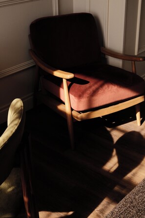 Una silla de madera con asiento y respaldo acolchados, ubicada en una habitación con piso alfombrado y una ventana que ilumina la silla.