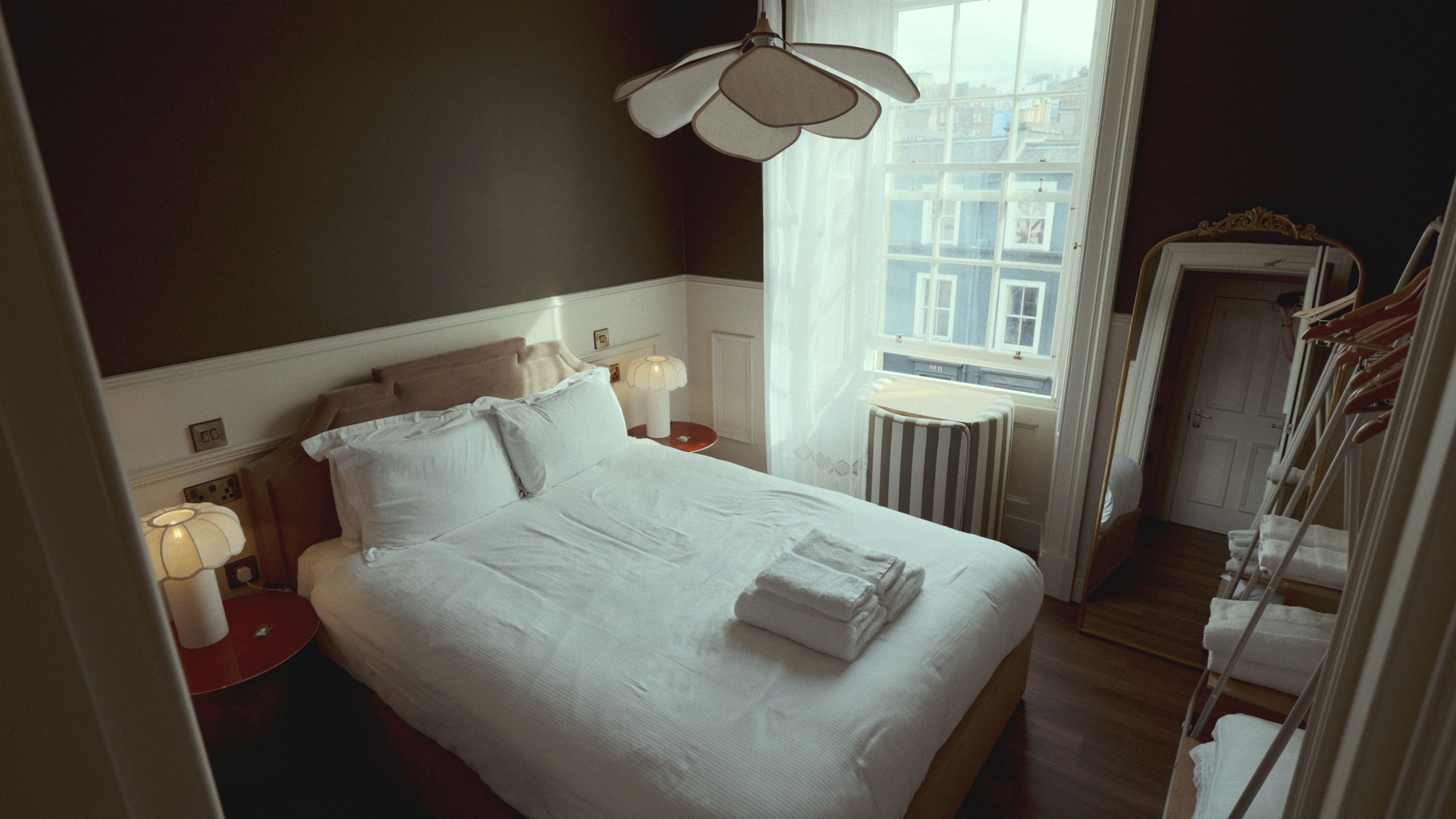 Habitación de hotel con una cama grande, lámparas de noche, vistas de la ciudad a través de la ventana y una puerta que da a un pasillo con un tendedero.