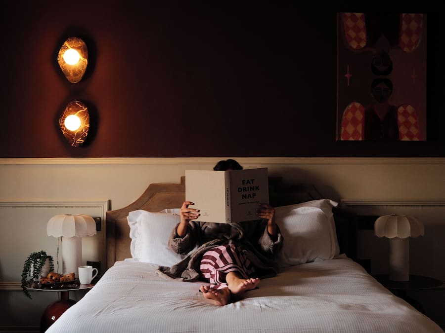 A person lying on a bed with a book titled 'EAT DRINK NAP' in a room with a dark wall and two wall-mounted lights.