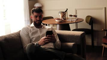 A man sitting on a couch using a smartphone in a room with a painting on the wall, a table with a pitcher and glasses, and a chair.