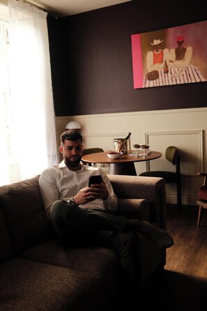 Un hombre sentado en un sofá usando un teléfono inteligente en una habitación con un cuadro en la pared, una mesa con una jarrón y vasos, y una silla.