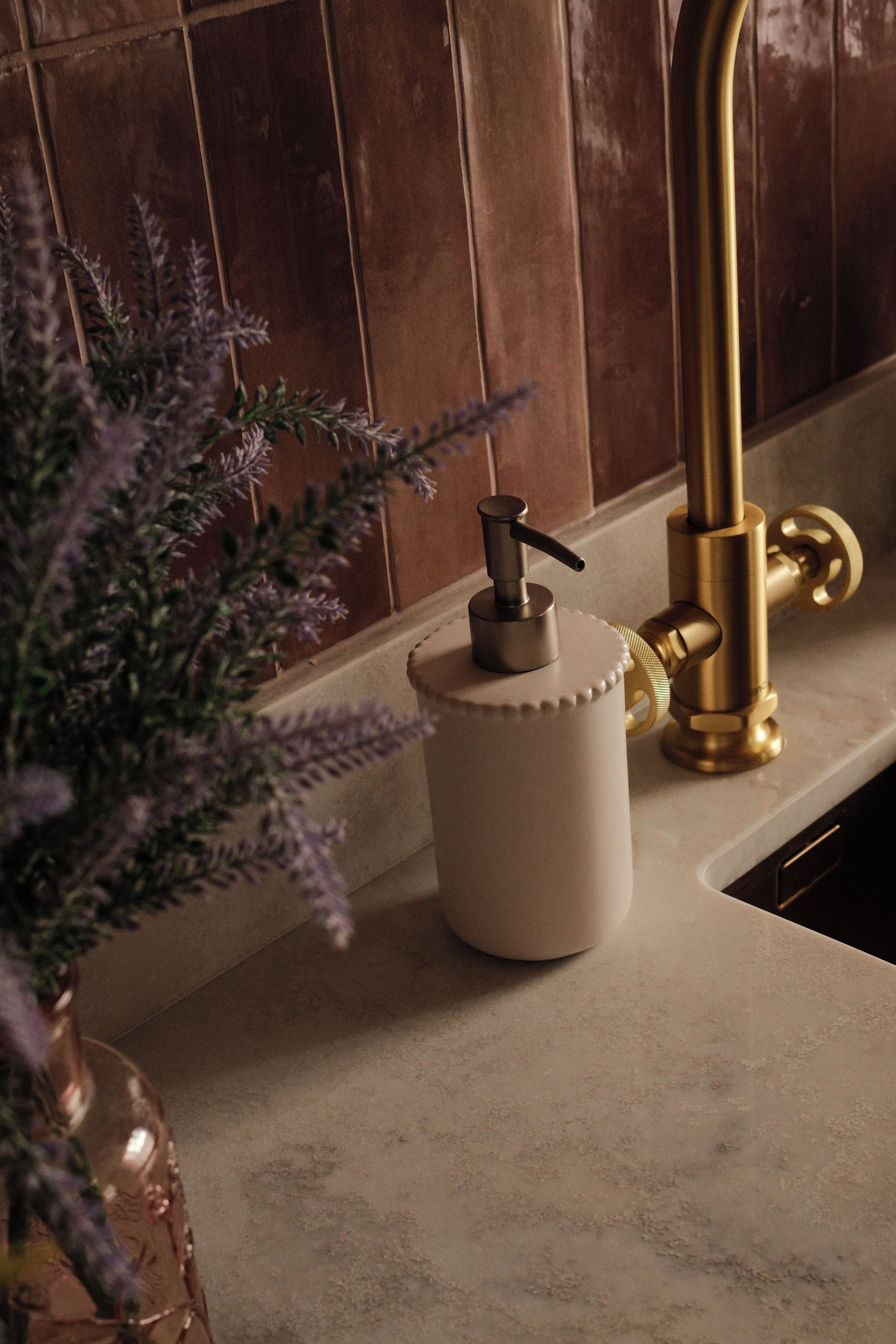 Un lavamanos de cocina con un grifo dorado, un dispensador de jabón y una maceta con flores moradas.