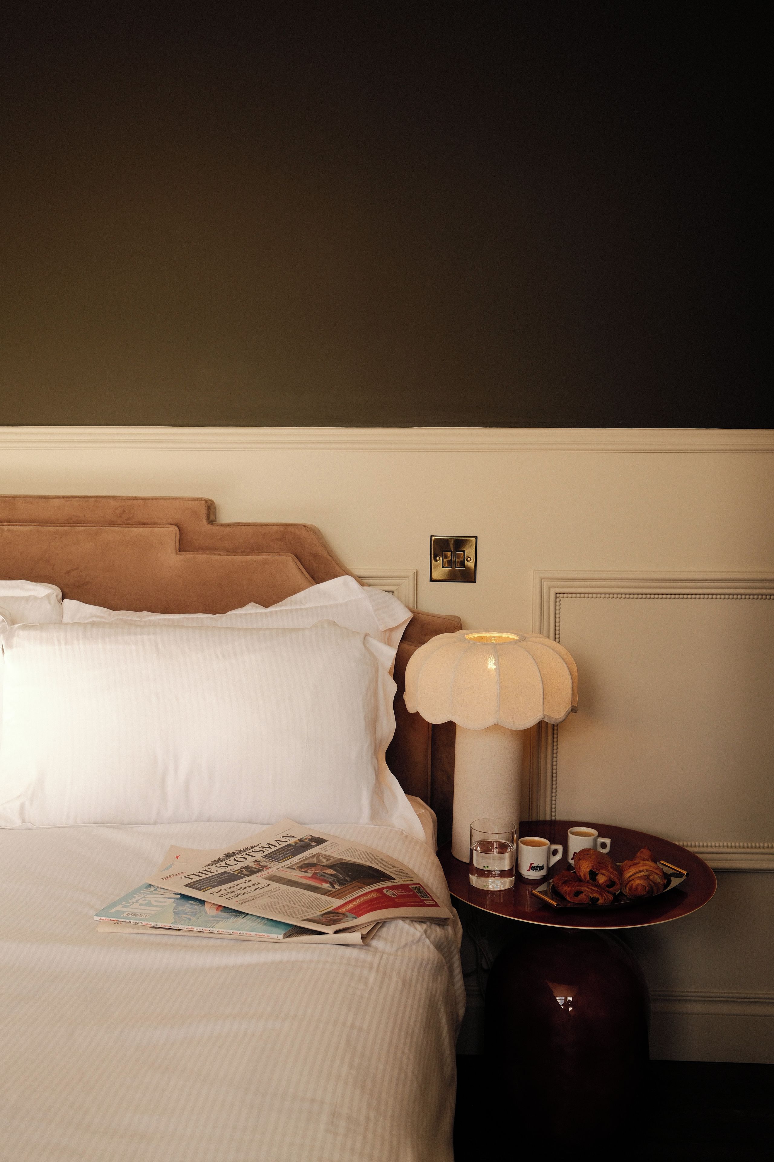Une chambre d’hôtel avec un lit, une table de chevet avec une lampe, un journal, un verre d’eau et un plateau avec des pâtisseries.