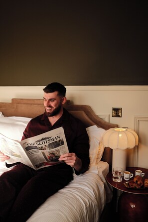 Una persona sentada en una cama leyendo un periódico, con una lámpara y un plato de croissants sobre una mesita de noche.