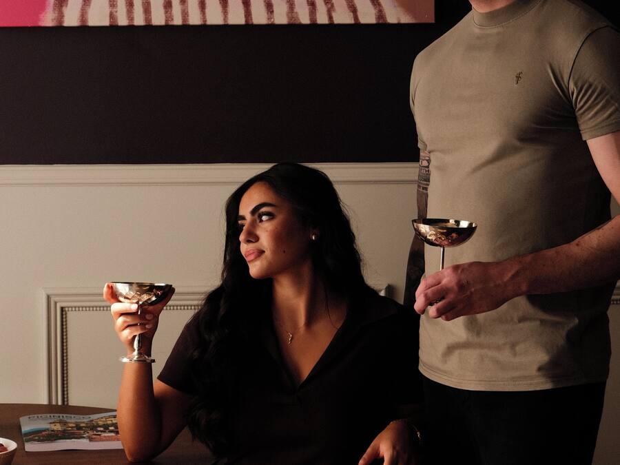A room with a painting on the wall, a table with a bottle and glasses, and a person holding a glass.