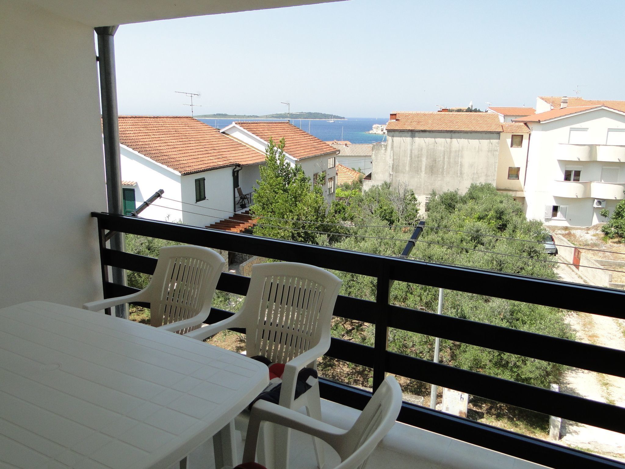 A balcony with a table and two chairs, overlooking a coastal area with buildings and a clear sky.