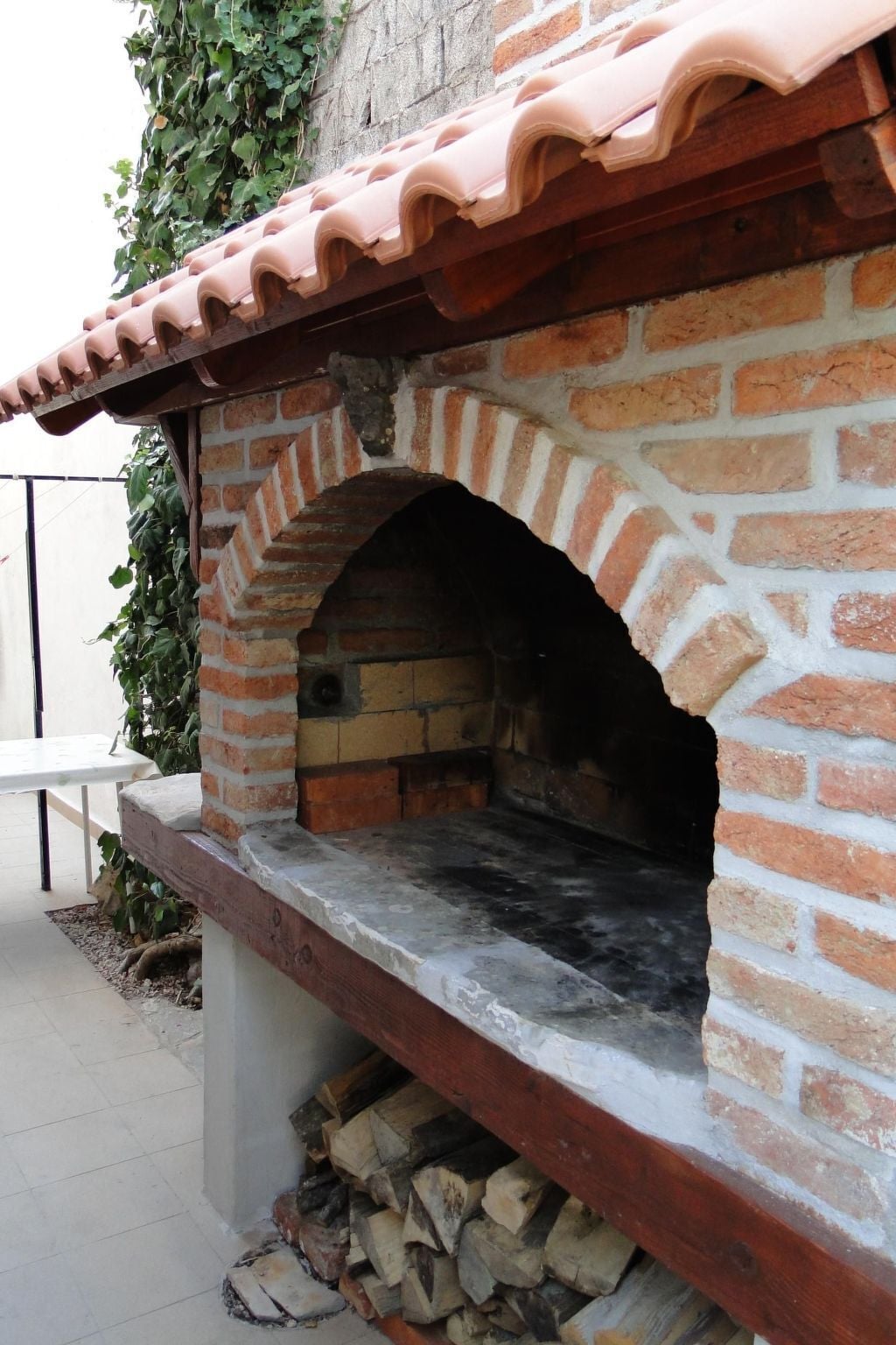 A brick-built outdoor oven with a terracotta roof, a stone bench, and a pile of firewood.