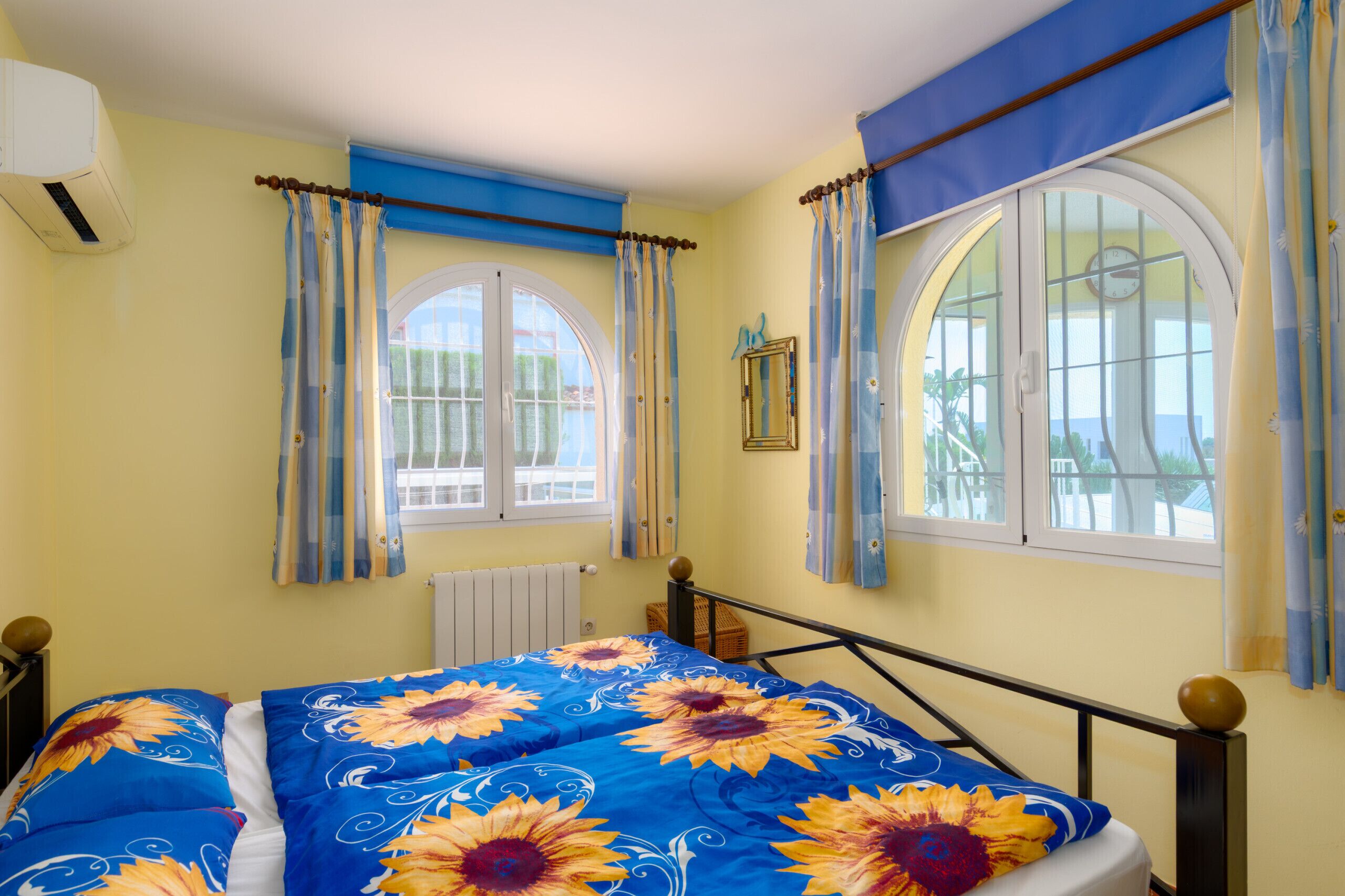A bedroom with a bed, two windows with curtains, a radiator, and a wall-mounted air conditioner.