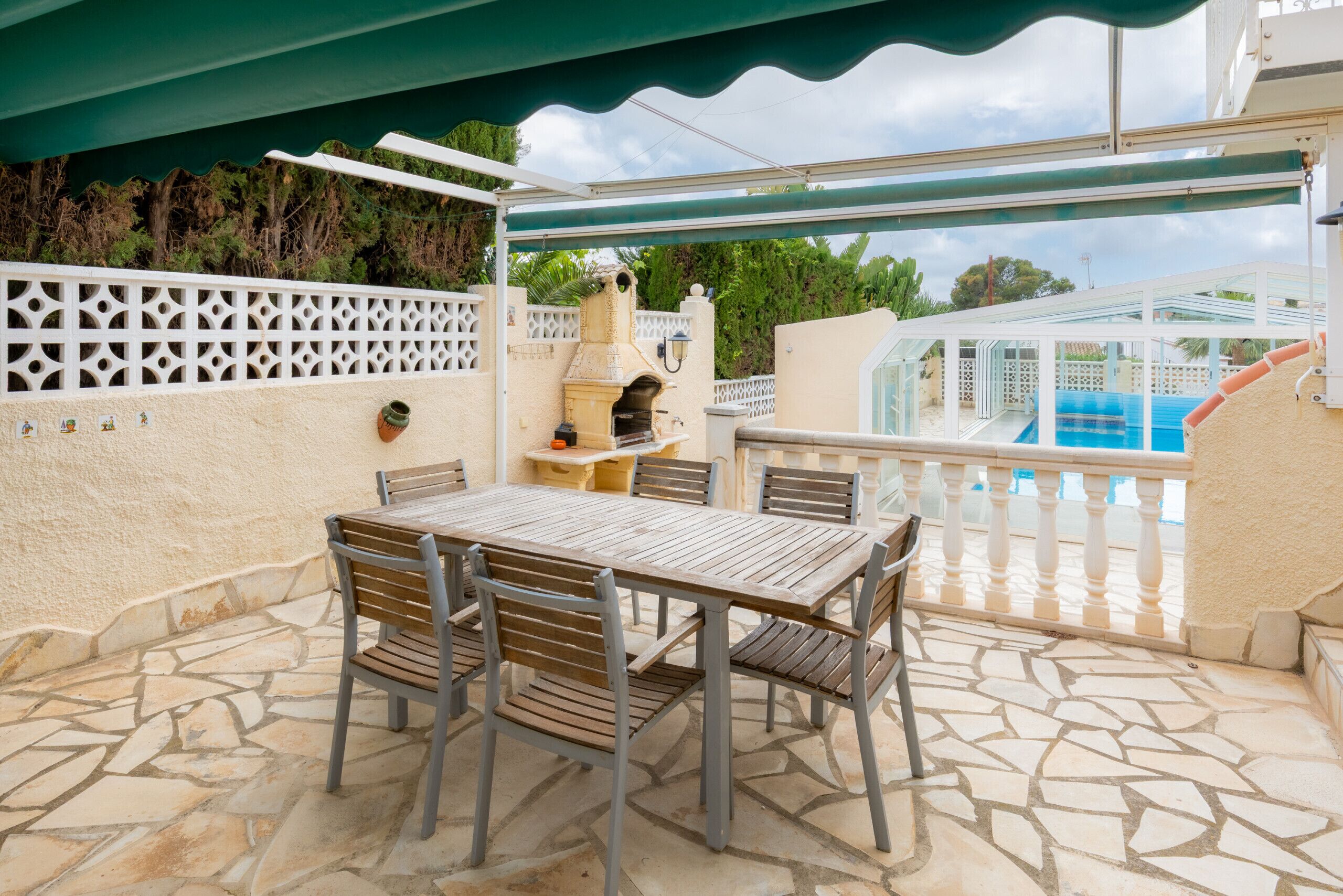 Un patio al aire libre con piso de piedra, una mesa de madera con sillas, una parrilla y una piscina al fondo.