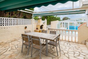 Eine Außenpatio mit Steinboden, einem Holztisch mit Stühlen, einem Grill und einem Pool im Hintergrund.