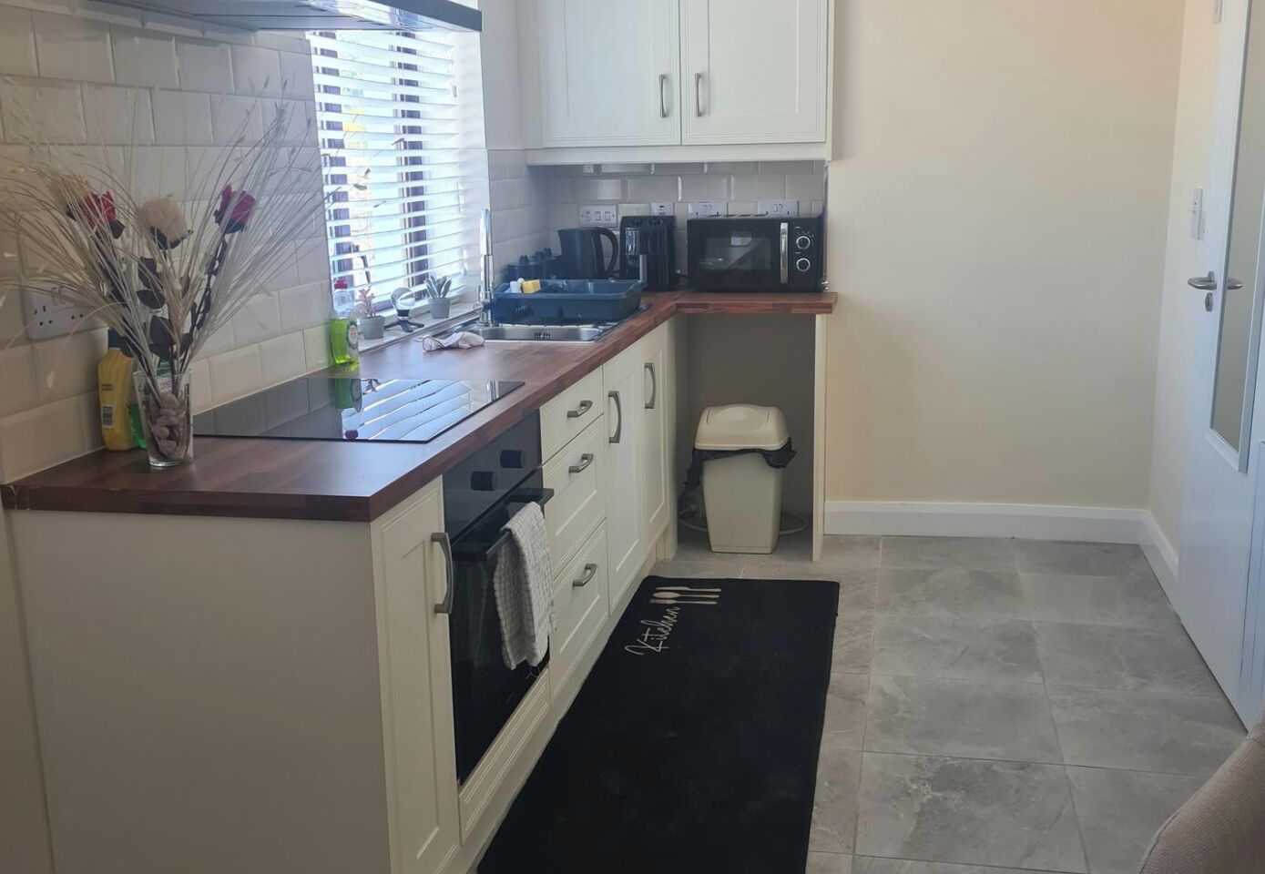 Cocina compacta con gabinetes blancos, microondas, lavamanos y un cubo de basura.