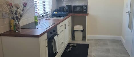 Cocina compacta con gabinetes blancos, microondas, lavamanos y un cubo de basura.