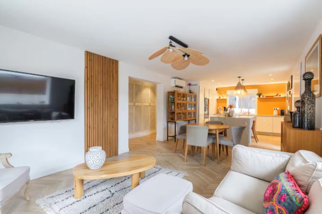 A modern living room with a flat-screen TV, a wooden accent wall, a dining area with a table and chairs, and a kitchen in the background.