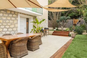 An outdoor patio with a wooden table and wicker chairs, a stone wall, and a grassy area with steps leading to a wooden deck. - Ground floor of a villa with private entrance and pool in Saint-Paul-de-Vence (Saint-Paul-de-Vence)