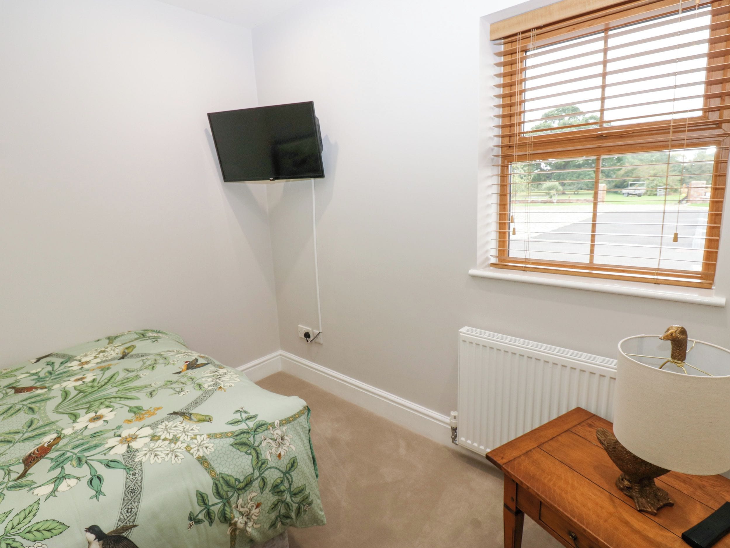 Une chambre avec un lit simple, un téléviseur fixé au mur, une fenêtre avec des stores, un radiateur et une table de chevet en bois avec une lampe.