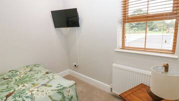 Une chambre avec un lit simple, un téléviseur fixé au mur, une fenêtre avec des stores, un radiateur et une table de chevet en bois avec une lampe.
