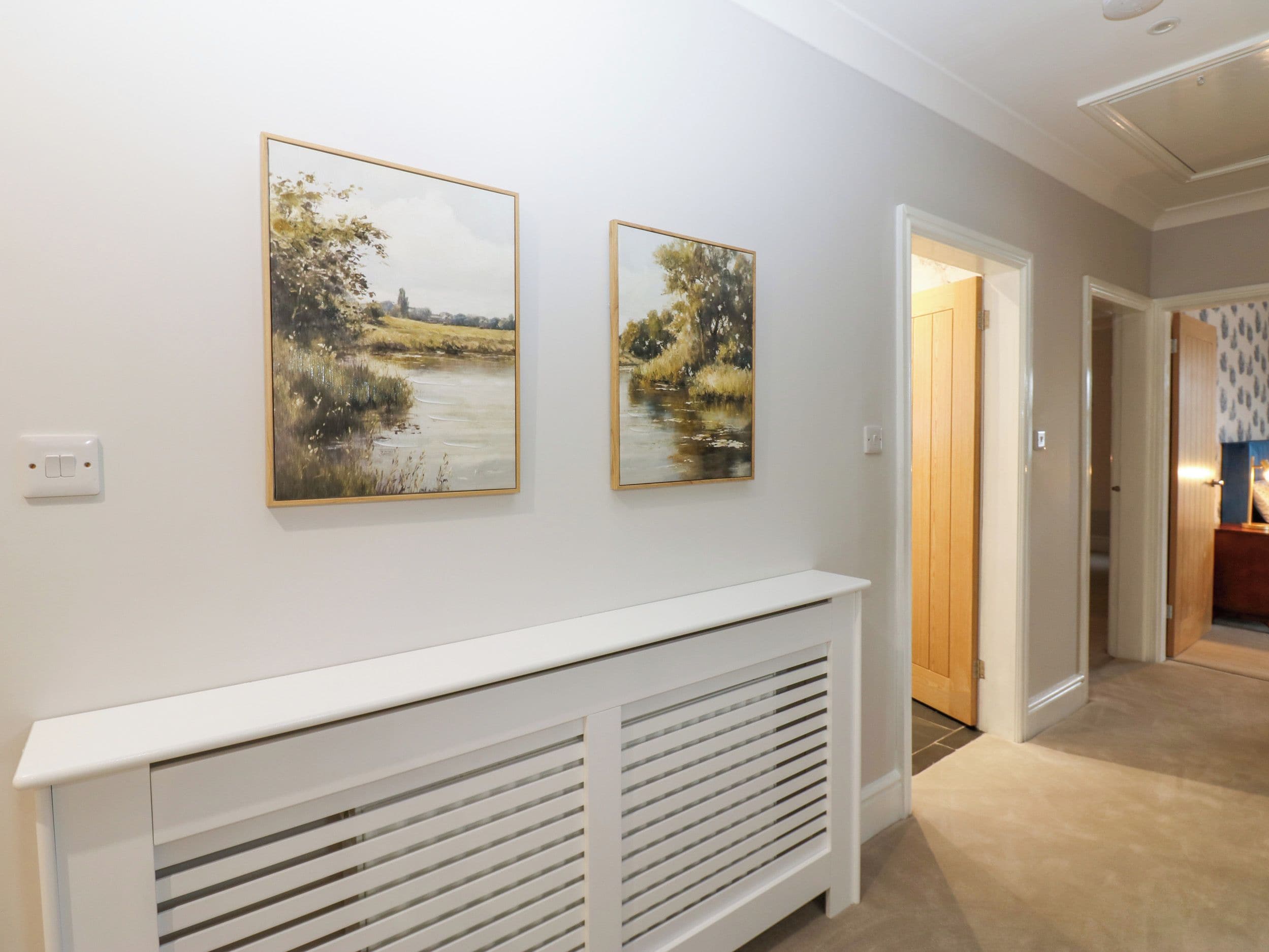 Um corredor com um radiador branco, duas pinturas de paisagens e uma porta que dá para outro quarto.