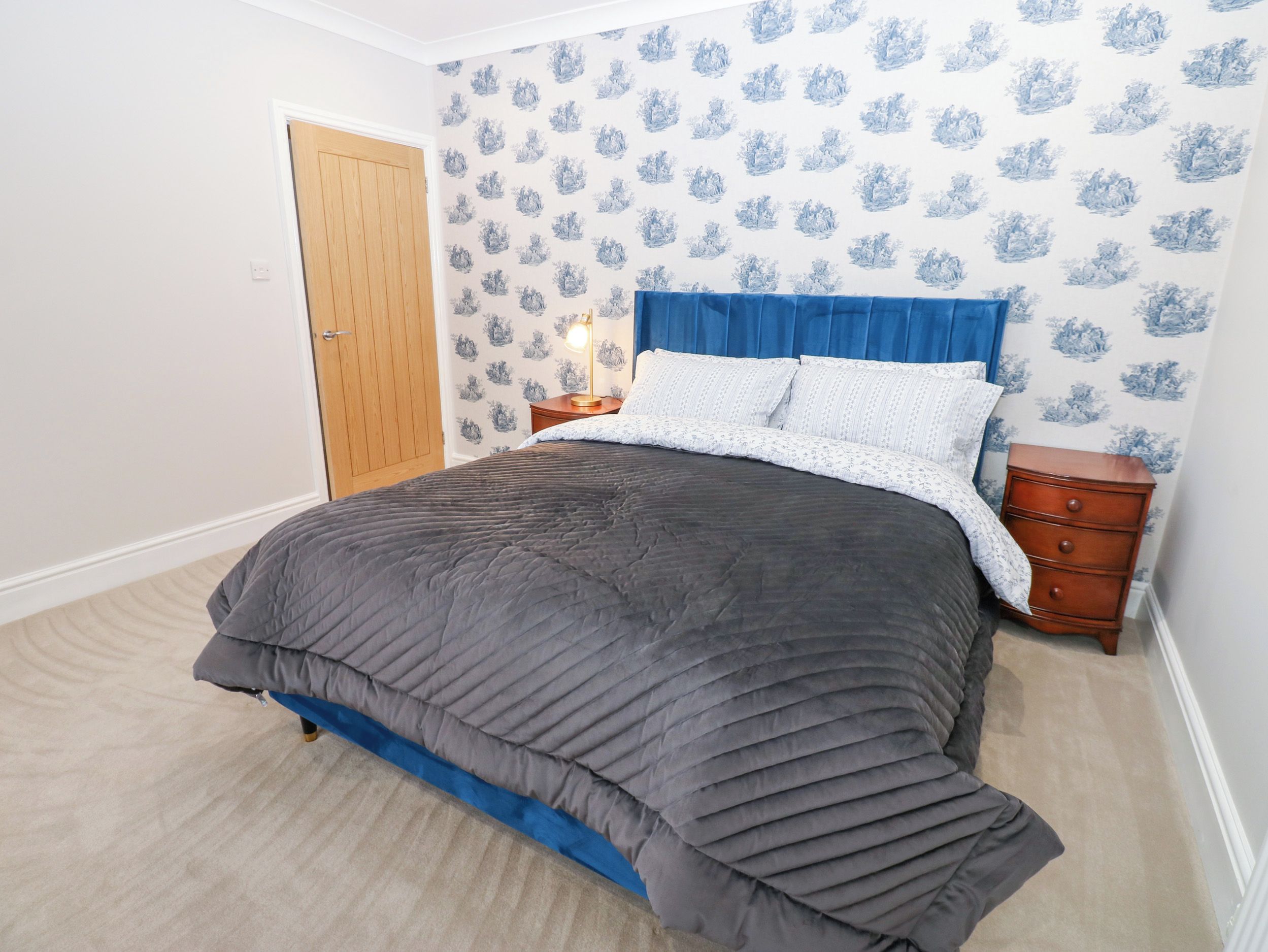 Une chambre à coucher avec un lit, une table de chevet, une commode, une porte et un papier peint à motifs.