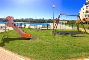 En lekplats med en rutschkana och gungor, en pool och en byggnad i bakgrunden.