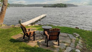 A lakeside seating area with two Adirondack chairs and a small round table, overlooking a calm lake with a dock extending into the water.