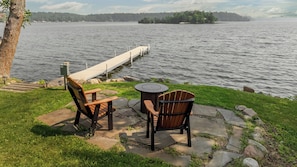 Un espacio de descanso junto al lago con dos sillas Adirondack y una pequeña mesa redonda, con vistas a un lago tranquilo y un muelle que se adentra en el agua.