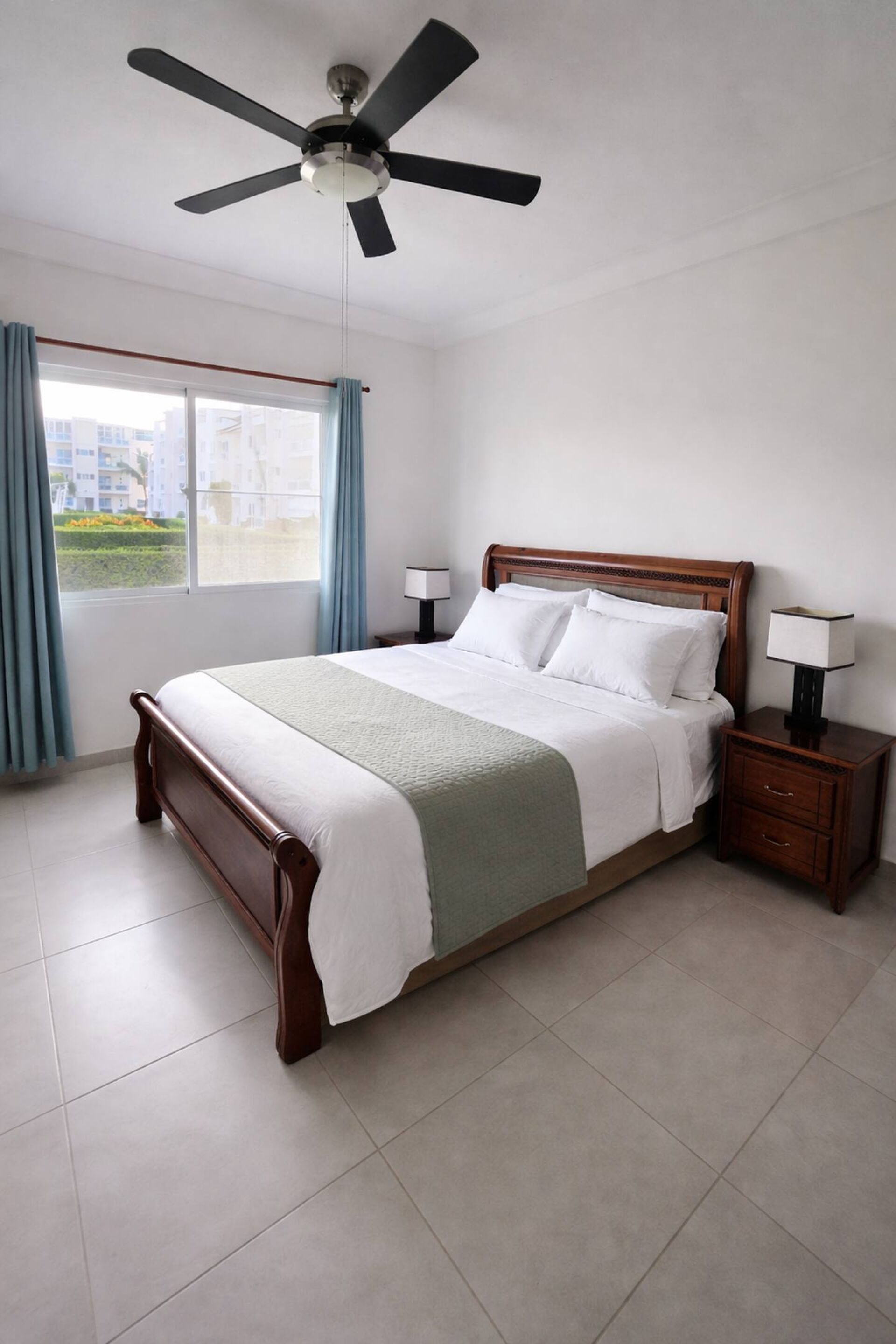 Habitación de hotel con cama, mesita de noche, ventilador de techo, ventana con cortinas y vista a edificios.