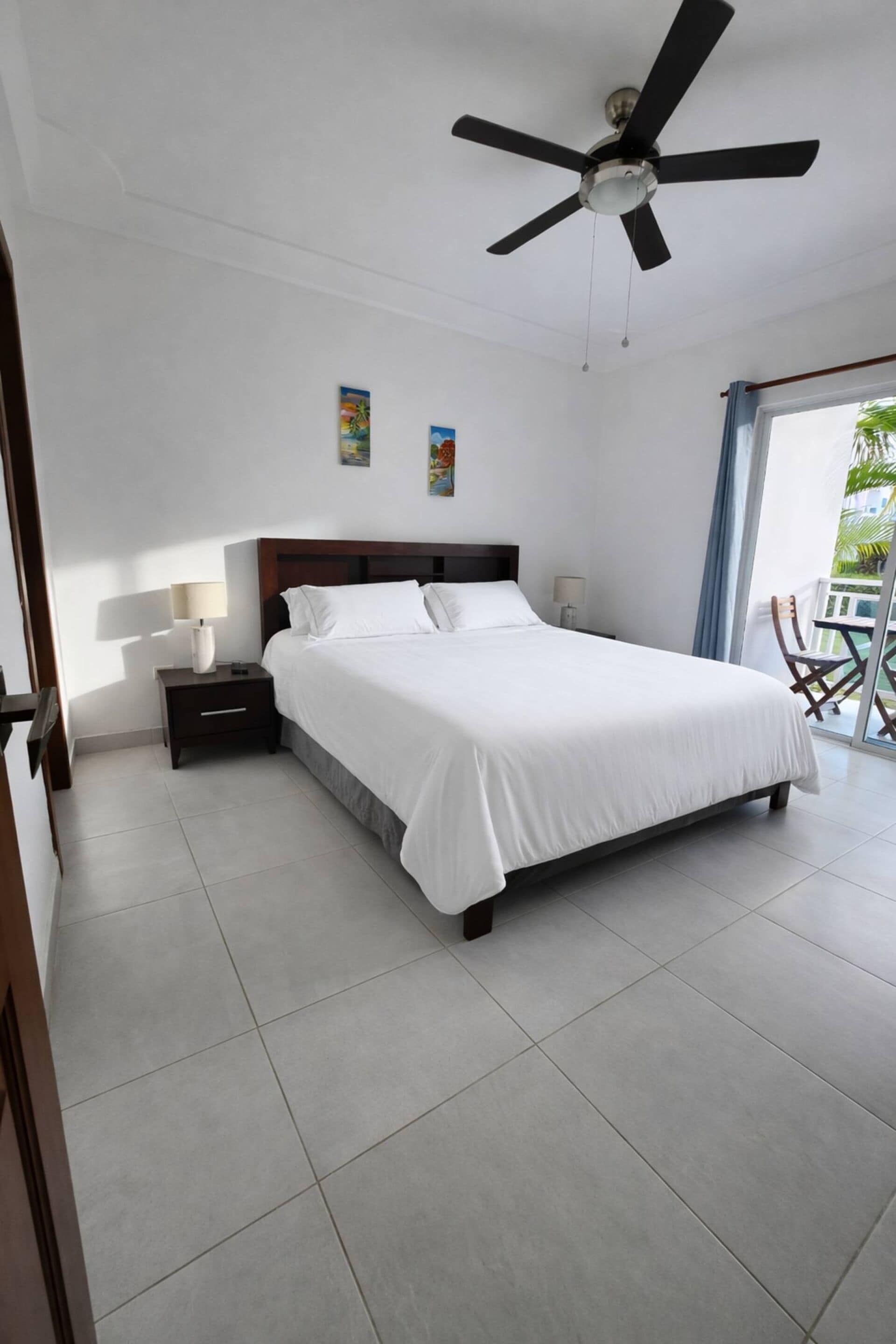 A hotel room with a bed, a nightstand, a ceiling fan, and a view of the outdoors.