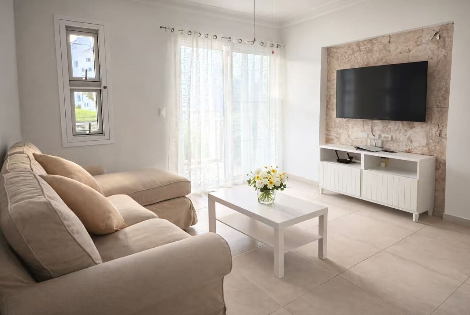 Un salon avec un canapé beige, une table basse blanche ornée d’un vase de fleurs, un téléviseur à écran plat fixé à un mur en marbre, et une fenêtre avec des rideaux légers.