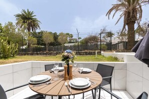 An outdoor dining area with a round table set for four, surrounded by chairs, with a view of a garden and a fence in the background. - Club Atlantis 150 - One Bed (Costa Adeje)