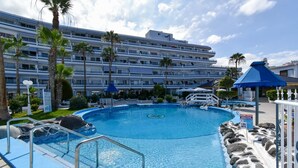 A swimming pool with a stone border, palm trees, and a hotel building in the background. - Club Atlantis 150 - One Bed (Costa Adeje)