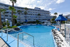 Ein Swimmingpool mit Steinrand, Palmen und einem Hotel im Hintergrund.