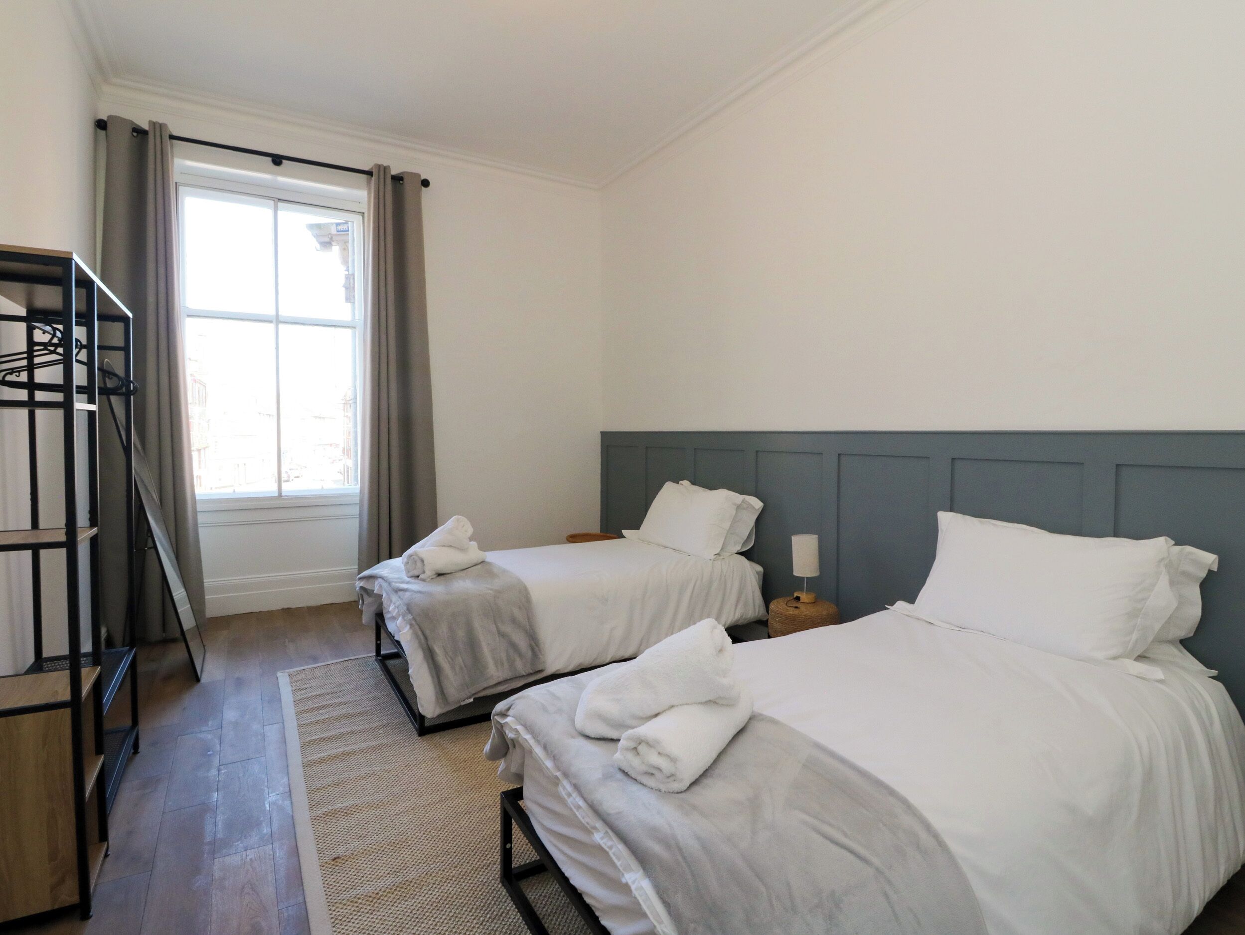 A hotel room with two beds, a window with curtains, a wooden floor, and a metal rack.