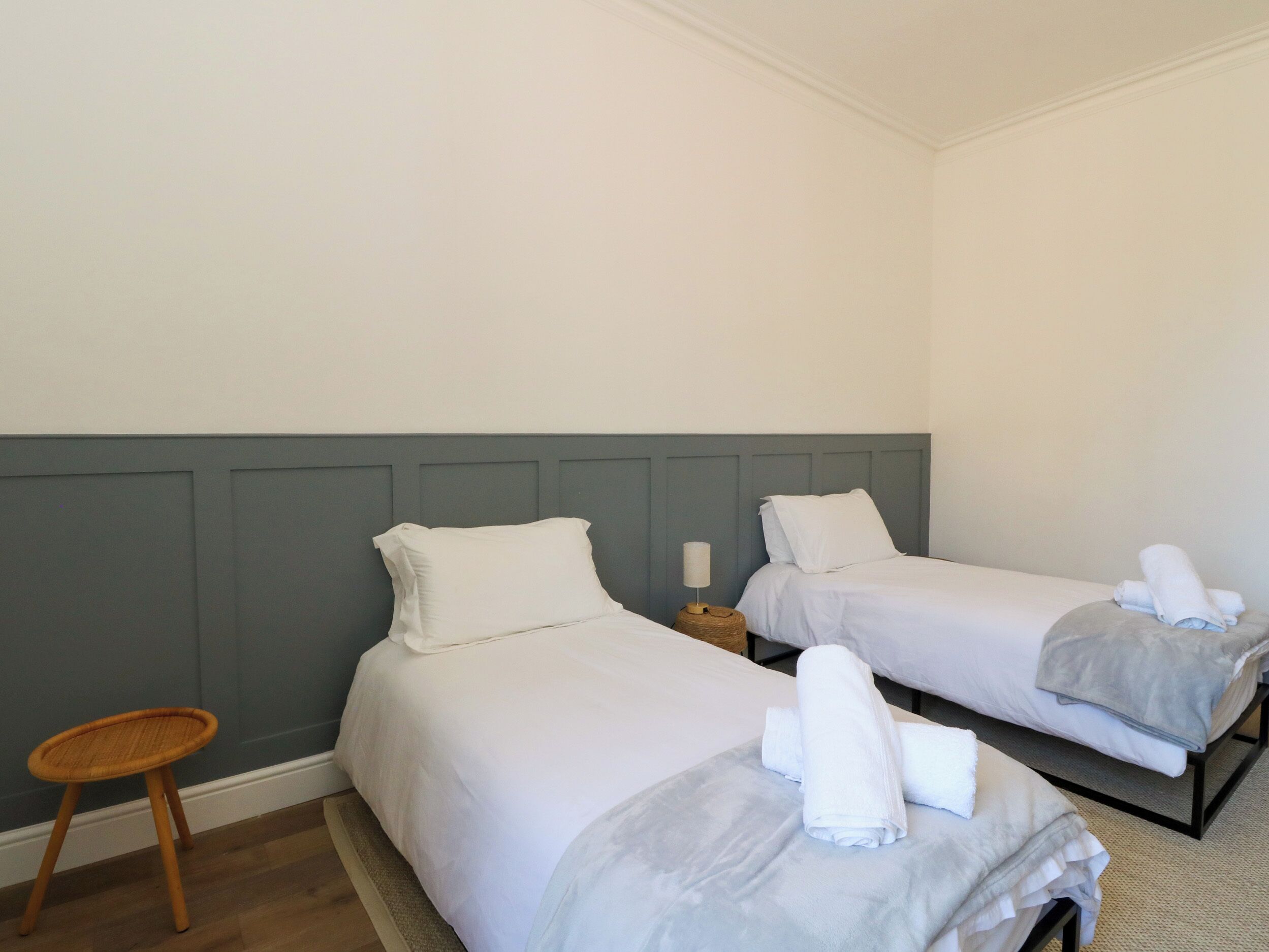 Una camera d'albergo con due letti singoli, un piccolo sgabello di legno e un comodino con una lampada.