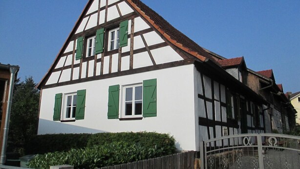 A traditional half-timbered house with a steep gable roof, a gated entrance, and a well-maintained hedge in front.