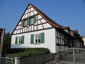 Une maison traditionnelle à colombages, dotée d’un toit à pignon abrupt, de volets en bois et d’une entrée fermée par un portail.