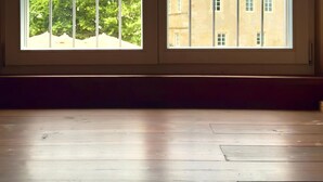 A room with a large window, wooden flooring, and a view of buildings and trees. - Schlossgut Franken (Redwitz an der Rodach)