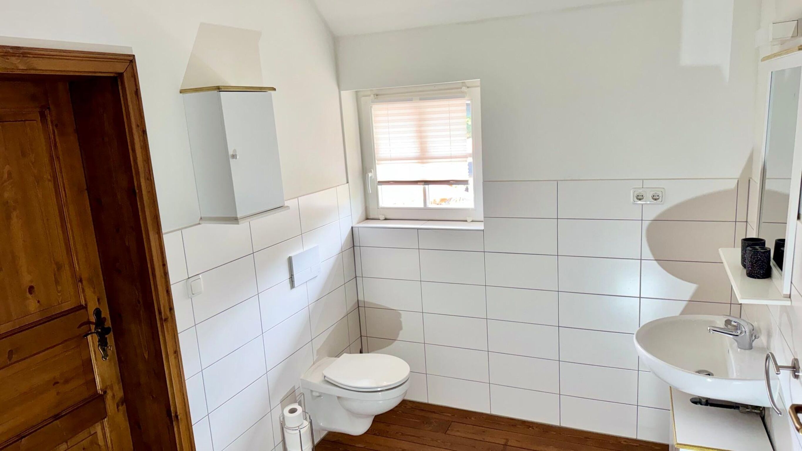 A bathroom with white tiles, a toilet, a sink, and a window.