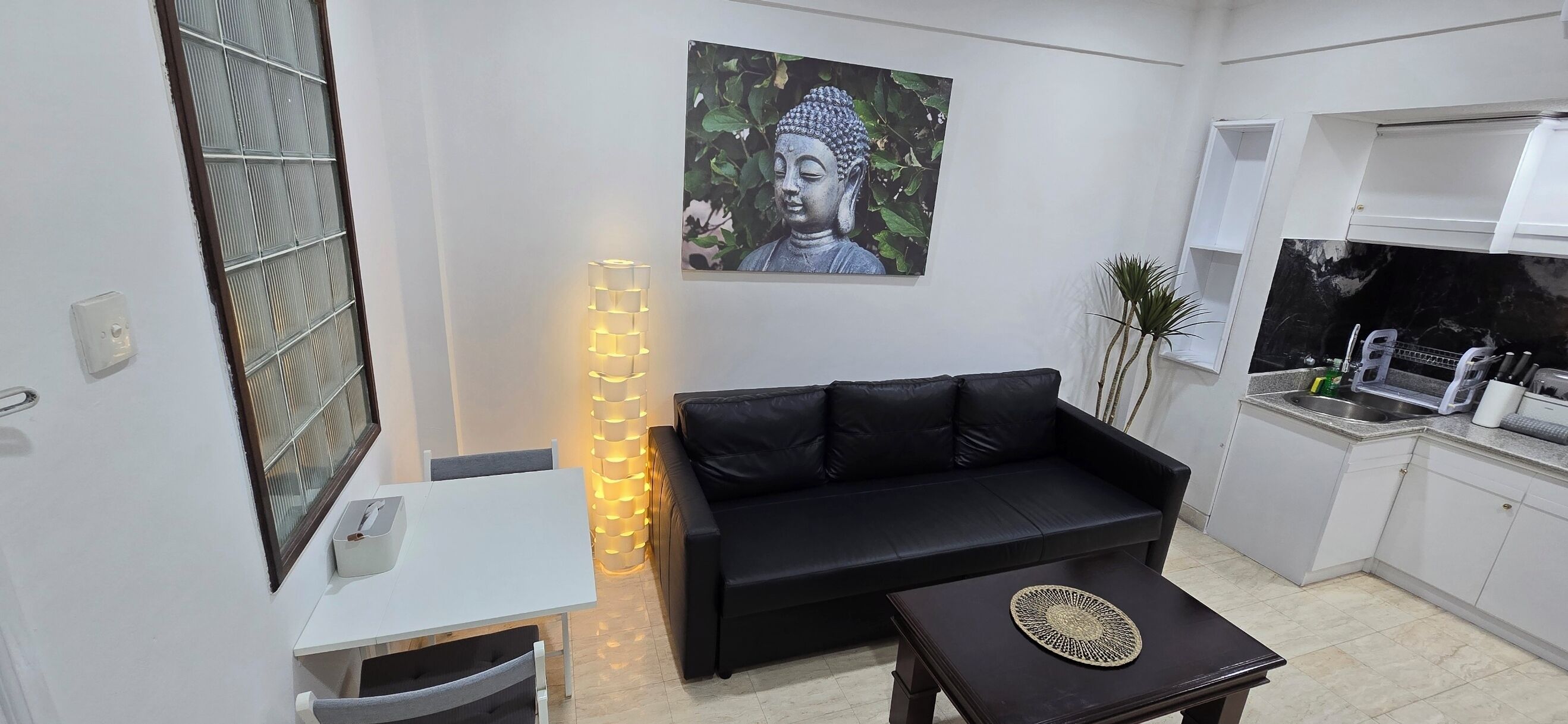 A compact living space with a black sofa, a small table, a wall-mounted Buddha statue, and a kitchen area with a sink and utensils.