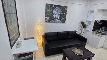 A compact living space with a black sofa, a small table, a wall-mounted Buddha statue, and a kitchen area with a sink and utensils.