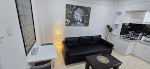 Un espacio habitable compacto con un sofá negro, una mesita, una estatua de Buda montada en la pared y una zona de cocina con un fregadero y utensilios.