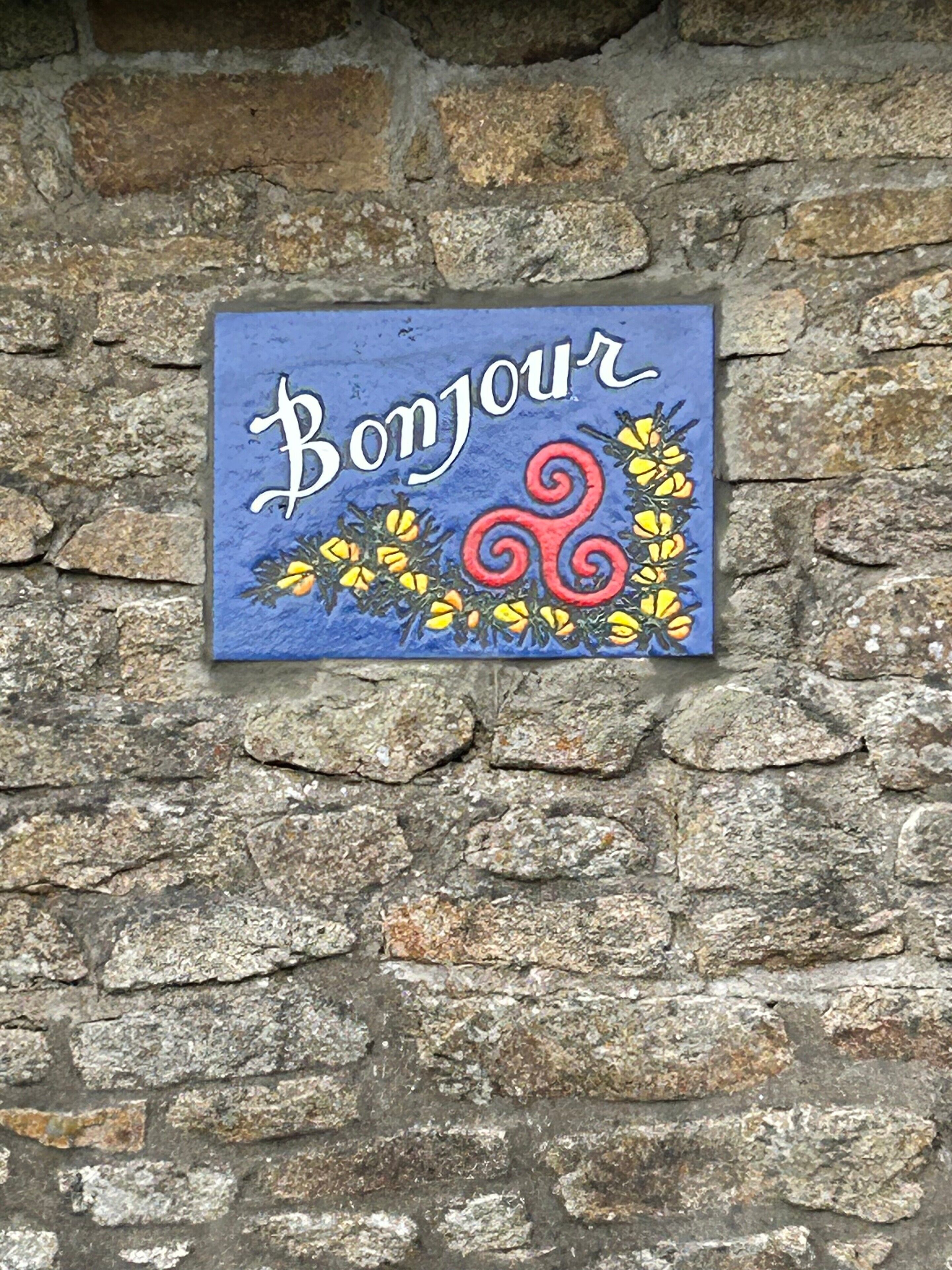 Uma placa azul com a palavra 'Bonjour' e elementos decorativos numa parede de pedra.