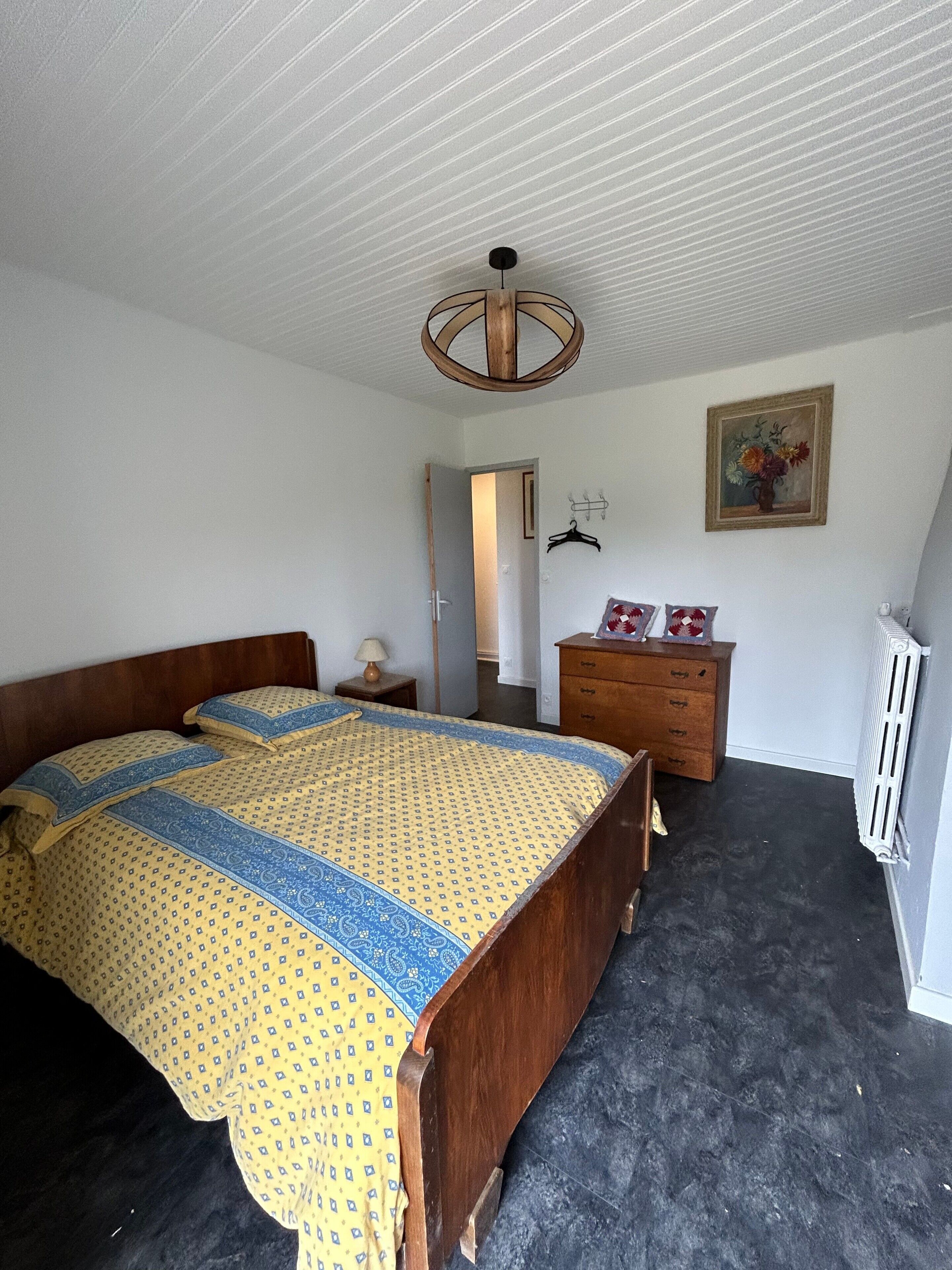 Un dormitori amb llit de fusta, un taulell, un radiador i una porta que dona a una altra habitació.