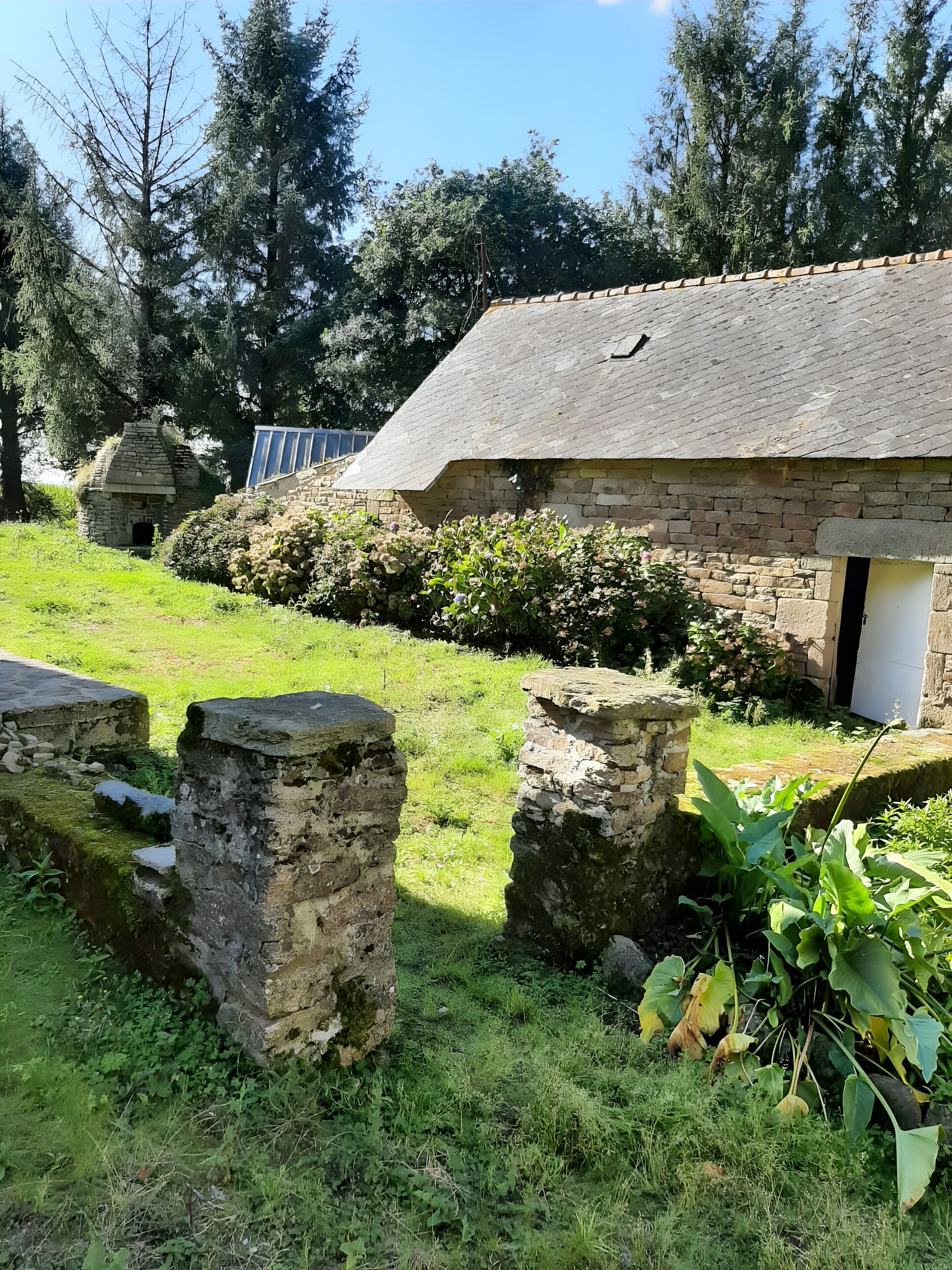 Un bâtiment en pierre, doté d’un toit en pente, entouré de verdure et d’un mur de pierre.